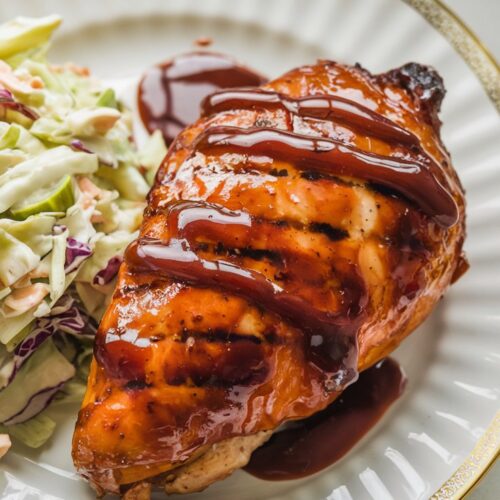 Glazed BBQ chicken breast with grill marks, served on a white plate with a side of coleslaw and a drizzle of extra BBQ sauce. The setting includes a checkered napkin on a wooden surface.