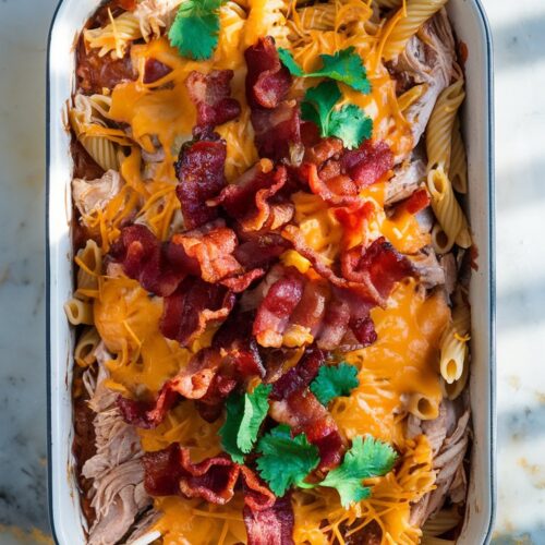 A white ceramic baking dish filled with tender shredded rotisserie chicken, cooked pasta, and tangy barbecue sauce, topped with crispy bacon and melted cheddar cheese. Garnished with fresh cilantro.