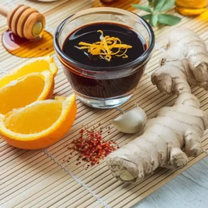 A glossy soy sauce captured in a clear glass bowl topped with orange shavings and surrounded by honey and fresh ginger and garlic.