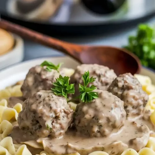 A plate of Swedish meatballs drenched in creamy gravy, served over buttered egg noodles, garnished with fresh parsley. A rustic wooden spoon resting on the side.