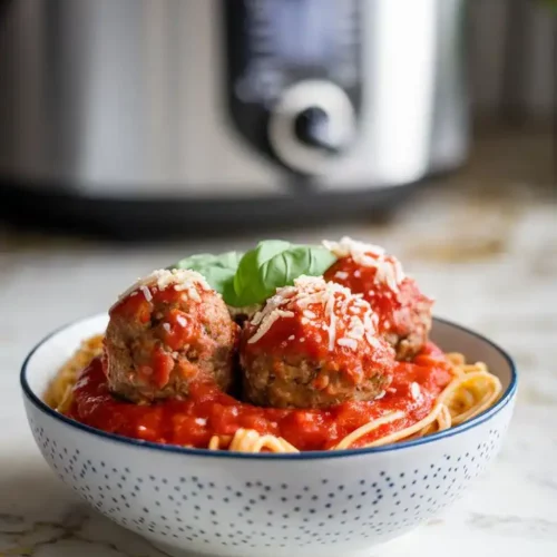 A bowl of tender meatballs smothered in rich marinara sauce, garnished with Parmesan and fresh basil, served with spaghetti. Shot on a white marble countertop with hints of gold and gray, in natural lighting.