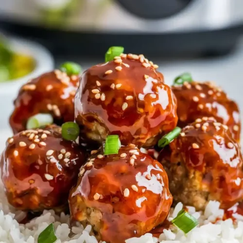 Glazed meatballs coated in a sticky honey garlic sauce, served over white rice, with sesame seeds and chopped green onions sprinkled on top.