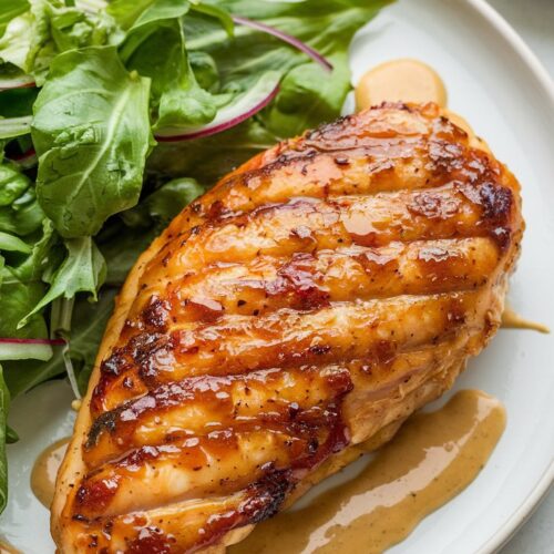 A caramelized honey mustard glazed chicken breast with a golden-brown exterior, served with a fresh green salad and a drizzle of honey mustard sauce on a white plate.