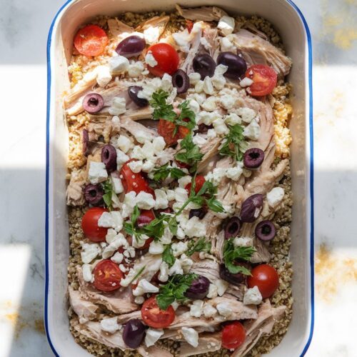 A white ceramic baking dish is filled with shredded rotisserie chicken, quinoa, diced tomatoes, kalamata olives, and crumbled feta cheese, with a light golden top. Garnished with fresh parsley.