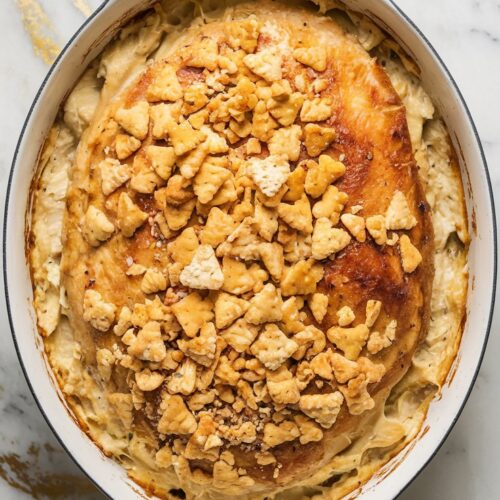 A creamy, golden-brown chicken casserole with a crunchy cracker topping, served in a white ceramic dish. The dish sits on a white marble counter with hints of gold and gray,