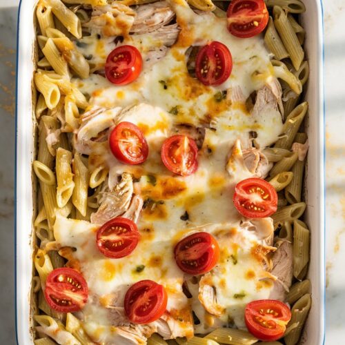 A white ceramic baking dish is filled with penne pasta coated in vibrant green basil pesto, shredded rotisserie chicken, and melted mozzarella, with cherry tomato halves scattered on top.