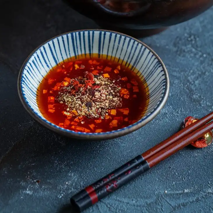 A fiery red Sichuan chili oil sauce in a small dipping bowl, topped with chili flakes and ground Sichuan peppercorns.