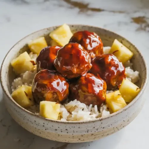 A bowl of glossy teriyaki meatballs coated in a rich, dark sauce, garnished with sesame seeds and green onions, with chunks of pineapple scattered around. Served over fluffy white rice in a ceramic bowl, set on a white marble countertop with hints of gold and gray, under soft natural lighting.