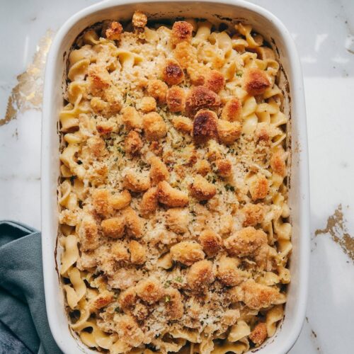 A close-up of a creamy tuna noodle casserole with golden brown breadcrumbs on top, served in a white ceramic baking dish. The dish sits on a white marble counter with hints of gold and gray, under natural lighting with soft shadows, taken in a top-down blogger-style shot.