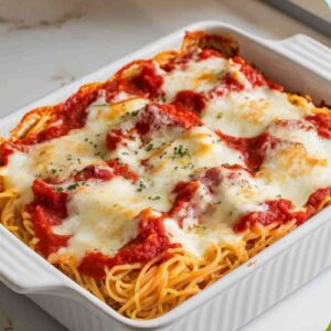 A gooey and indulgent Million Dollar Spaghetti bake with layers of creamy cheese, marinara sauce, and melted mozzarella on top, served in a white ceramic baking dish. The dish sits on a white marble counter with hints of gold and gray, under soft natural lighting, taken from a top-down perspective.