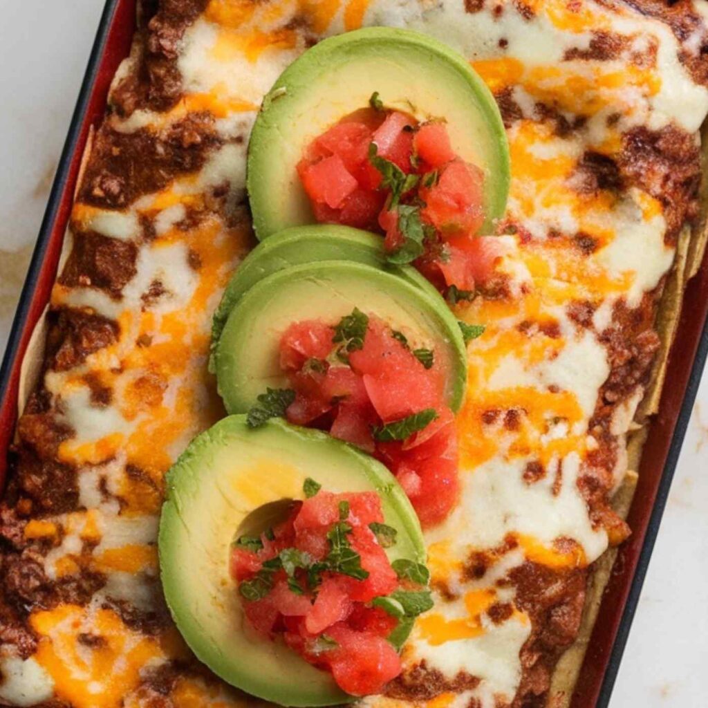 red casserole dish with ground beef and melted cheese on top garnished with avocado slices and tomatoes
