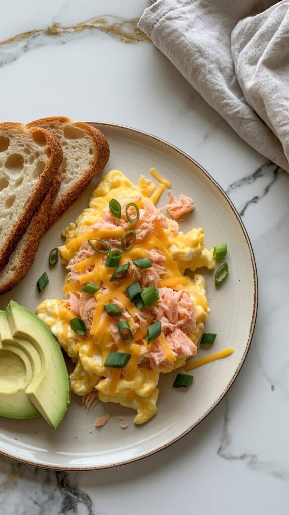fluffy scrambled eggs m ixed with canned salmon and green onions scattered on top, avocado slices and toaste on the side