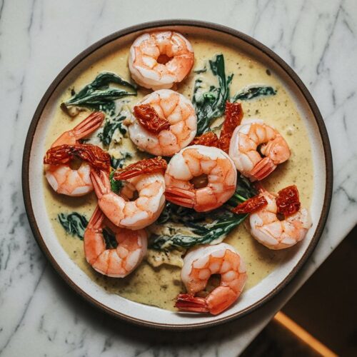 A white plate showcasing luxurious garlic-Parmesan cream sauce enveloping plump, pink shrimp, garnished with sun-dried tomatoes and fresh spinach.