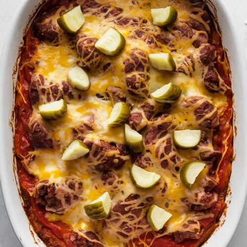 A white casserole dish containing a hearty dish. The dish appears to be a baked casserole with a sweet-savory tomato sauce and melted cheddar, melted cheese on top, and garnished with diced pickles, shown from above with soft lighting.