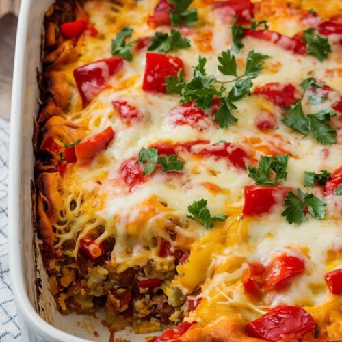 A white casserole dish containing a hearty dish. The dish appears to be a baked casserole with melted cheese and colorful chopped peppers, melted cheese on top, and garnished with parsley flakes, captured from the side to show the cheese texture.