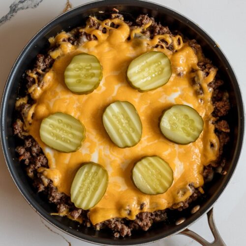 A cheesy ground beef skillet topped with melted cheddar and scattered pickle pieces,