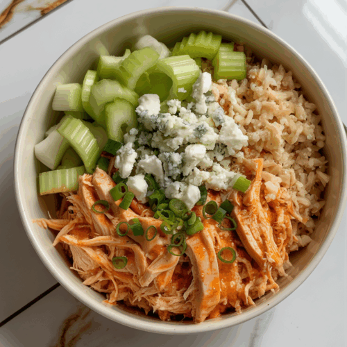 A spicy buffalo chicken bowl with shredded chicken, cauliflower rice, celery, crumbled blue cheese, and green onions, served in a white bowl on a white countertop with grey and gold veining.