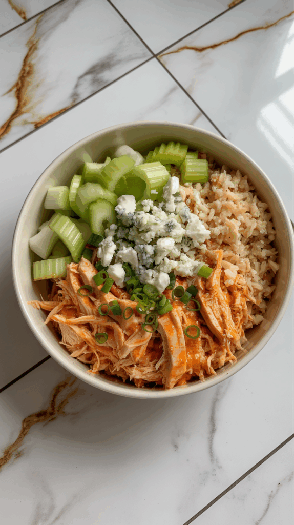 A spicy buffalo chicken bowl with shredded chicken, cauliflower rice, celery, crumbled blue cheese, and green onions, served in a white bowl on a white countertop with grey and gold veining.