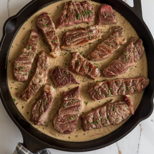 A cast iron skillet filled with seared ribeye strips in creamy sauce