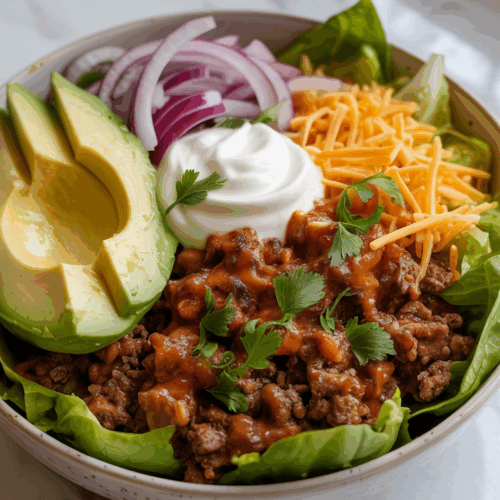 A hearty low carb taco bowl with seasoned ground beef, shredded cheddar, lettuce, diced avocado, red onion, and sour cream, served in a white bowl on a white countertop with grey and gold veining.