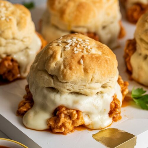 **Alt Text for Image:** Golden brown Chicken Biscuit Bombs resting on a rustic wooden board. Each biscuit bomb is flaky and buttery, filled with a savory blend of ground chicken mixed with barbecue sauce and melted mozzarella. The tops are sprinkled with sesame seeds, adding a touch of crunch. Fresh parsley is scattered around the board, enhancing the visual appeal. Accompanying the bombs is a small bowl of barbecue sauce for dipping, set against a cozy kitchen backdrop.