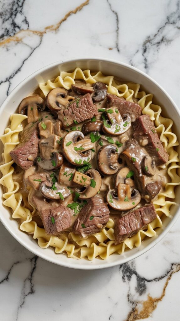 A shallow white bowl of creamy beef stroganoff made with sirloin strips, mushrooms, and egg noodles, topped with chopped parsley. The sauce is rich and glossy, clinging to the noodles and beef