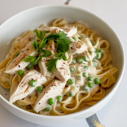 A white bowl of crockpot chicken Alfredo with green peas, served over pasta and topped with chopped parsley.