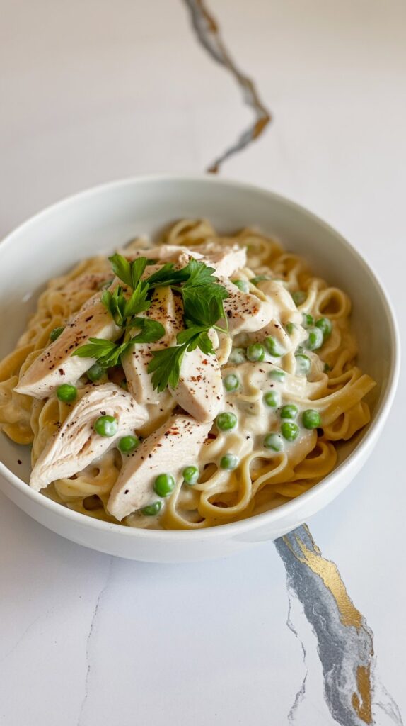A white bowl of crockpot chicken Alfredo with green peas, served over pasta and topped with chopped parsley.