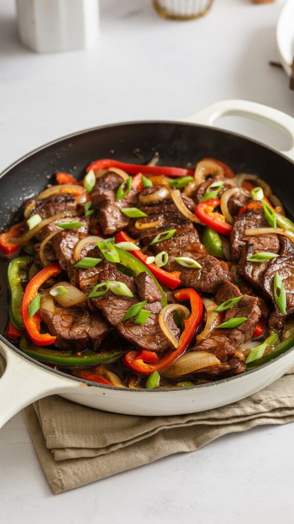 a skillet with strips of steak and red peppers also onions and scallions