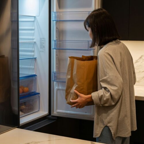 A person staring blankly into an open fridge with almost nothing inside, holding a half-empty grocery bag. Disorganized counter nearby with a few random items.