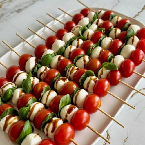 A white platter filled with Caprese skewers made of cherry tomatoes, basil leaves, and mozzarella balls, arranged in neat rows. Drizzled with balsamic glaze.