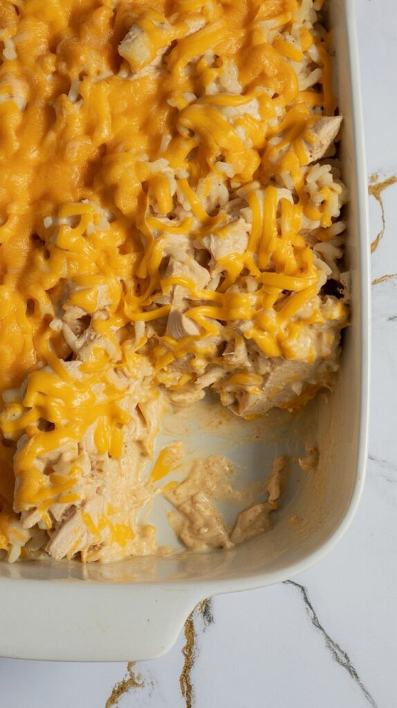 Chopped chicken and rice baked in a creamy cheddar sauce, topped with melted cheese. Presented in a white casserole dish on a clean white and gold-veined counter. Casserole is uncut.