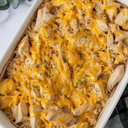 Creamy baked casserole with chopped chicken, melted cheddar cheese, and golden cracker topping in a white dish on a white countertop with gray and gold veining. Casserole is uncut and untouched.