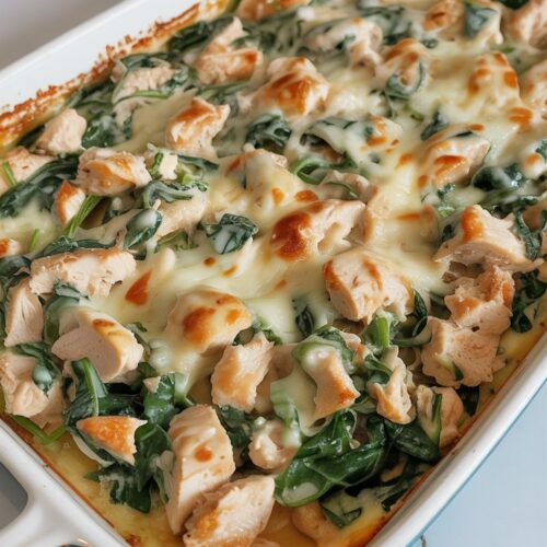 Chopped chicken and spinach in a creamy base with melted cheese, shown in a white casserole dish on a gray-veined white countertop. Uncut and neatly presented.