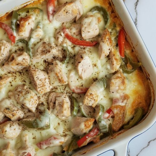 Casserole with chopped chicken, bell peppers, onions, and provolone cheese, shown in a white baking dish on a clean quartz counter. Dish is unsliced and intact.