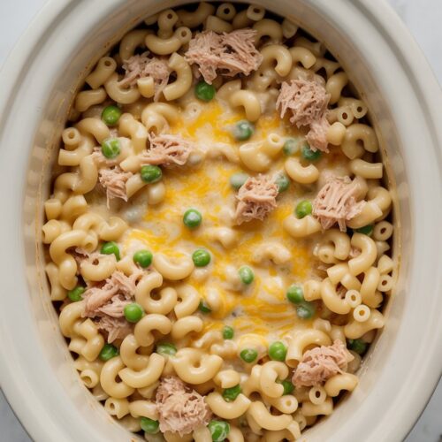A crockpot insert filled with creamy tuna noodle casserole made with elbow macaroni, peas, and melted cheese, viewed from above on a clean white countertop.