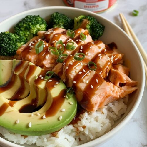 a bowl of white rice topped with flaked salmon from a can and a slice of avocado with teriyaki sauced on top. A can of salmon in the background