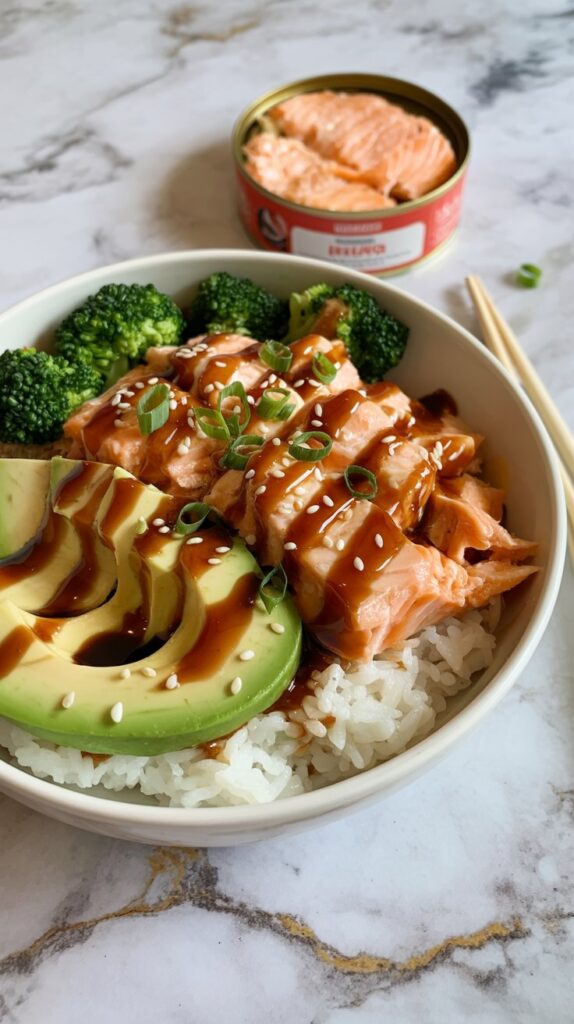 a bowl of white rice topped with flaked salmon from a can and a slice of avocado with teriyaki sauced on top. A can of salmon in the background