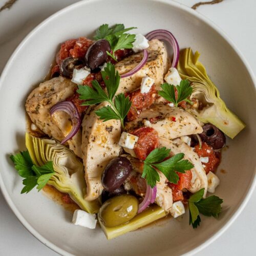 Mediterranean chicken with artichokes, olives, red onion, and feta in a shallow white bowl