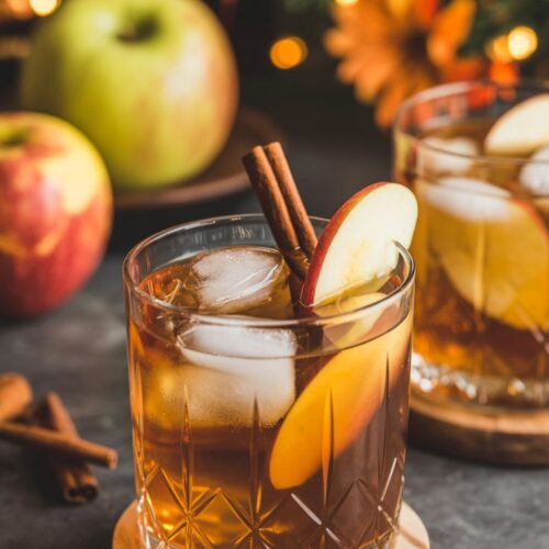 A glass filled with amber Apple Cider Bourbon Fizz cocktail, served over ice in a rocks glass, garnished with an apple slice and cinnamon stick, with fall decor in the background