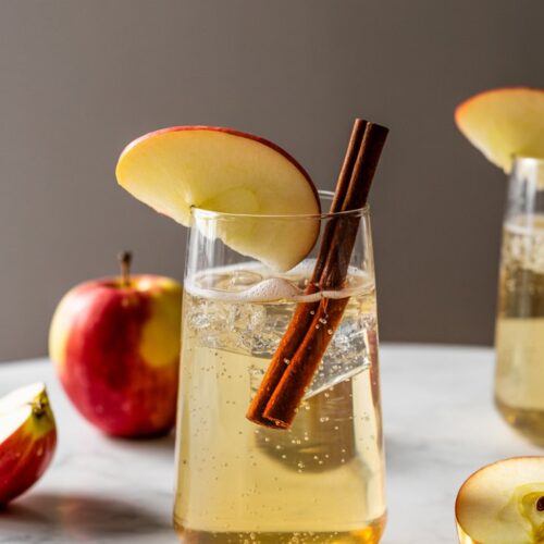 Sparkling apple and ginger mocktail in a stemless glass, garnished with a cinnamon stick and apple slice, styled with autumn colors and textures.