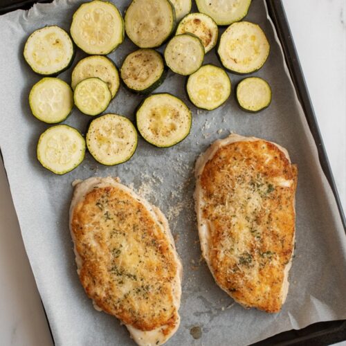 two parmesan-crusted chicken cutlets and roasted zucchini sliced into ¼-inch rounds or half-moons, on a parchment-lined sheet pan, placed on a white quartz countertop
