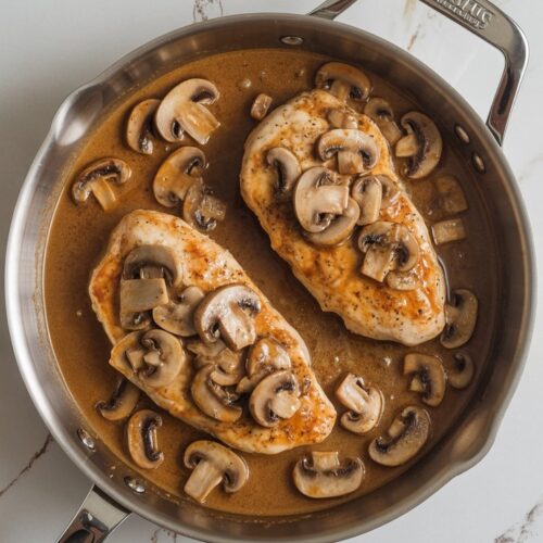 Chicken marsala with golden chicken cutlets and mushrooms in sauce, served in a stainless skillet on a white quartz countertop with grey and gold veining.