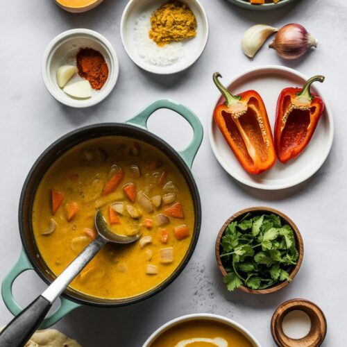 A top view of ingredients to make curry butternut squash soup including squash cut in half, spices, a bowl of finished soup and a dutch oven of the soup with immersion blender immersed.
