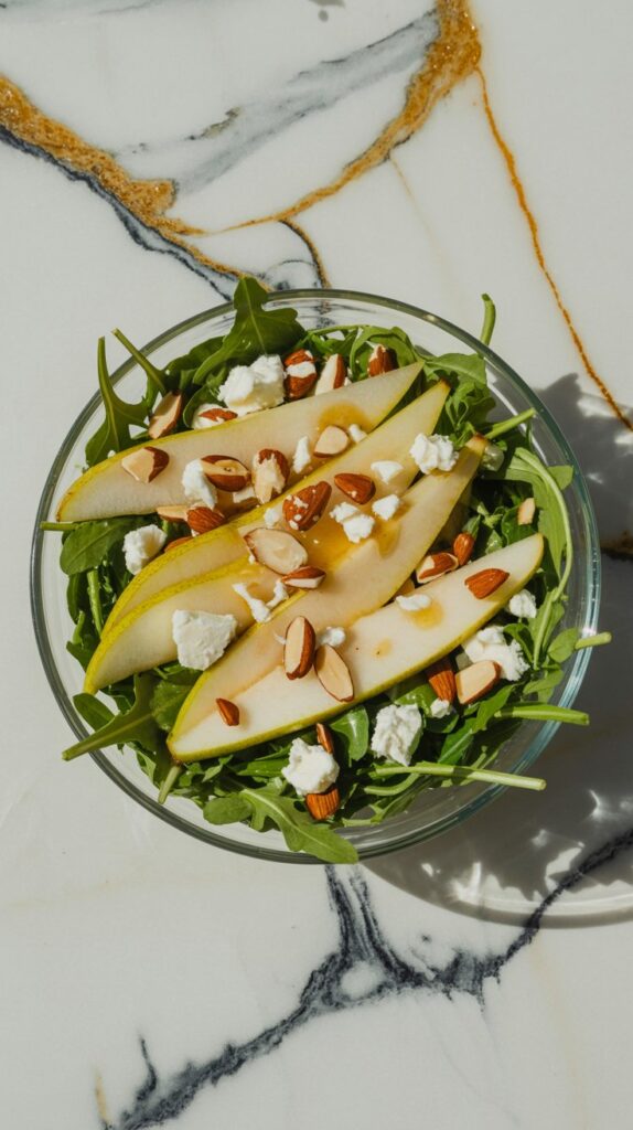 Overhead photo of arugula salad with pear slices, goat cheese, and almonds in a glass bowl.