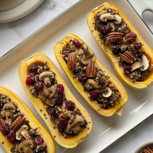 Overhead view of a white serving platter with roasted delicata squash halves stuffed with quinoa, mushrooms, cranberries, and pecans, centered on a white quartz countertop.