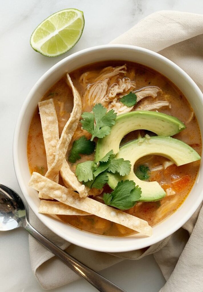 A bowl of rich brothy soup with slices of avocado, shredded chicken and toasted tortilla strips.