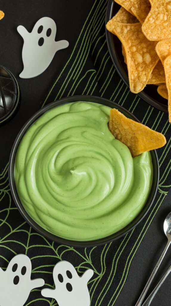 Green salsa verde and sour cream swirled together in a black bowl to resemble bubbling potion, with ghost-shaped tortilla chips on the side.