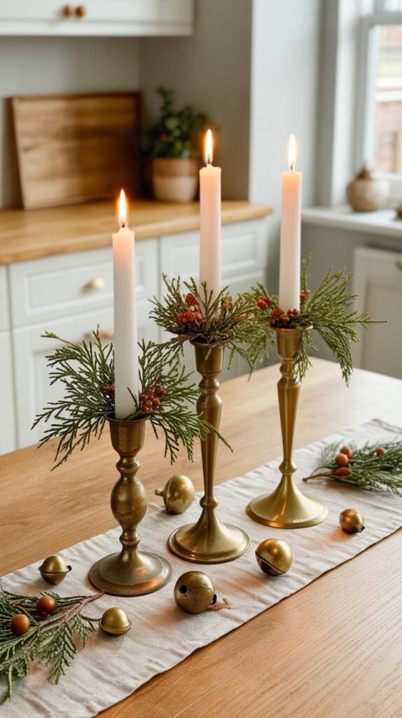 Brass candlesticks of varying heights with white tapers surrounded by pine sprigs and rustic bells, styled as a cozy Christmas table centerpiece.