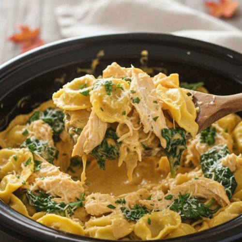 Slow cooker of cheese tortellini with shredded chicken and spinach in creamy pumpkin Alfredo sauce, a wooden spoon lifting a scoop